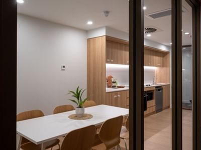 Modern hotel kitchen and dining area with wooden cabinets, white table, and potted plant at UniLodge Brisbane City.