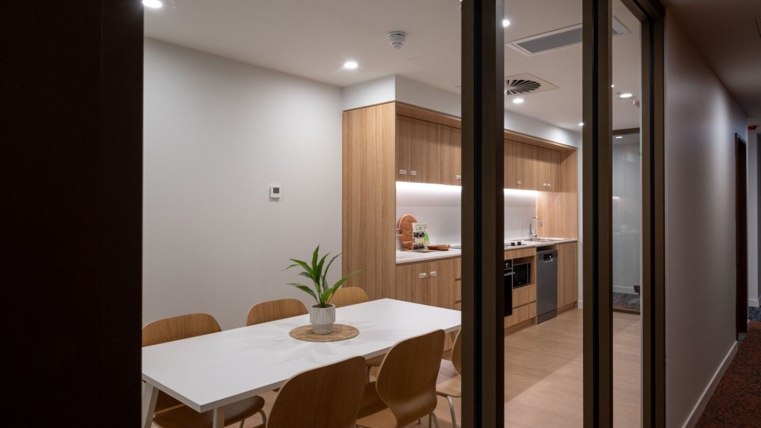 Modern hotel kitchen and dining area with wooden cabinets, white table, and potted plant at UniLodge Brisbane City.