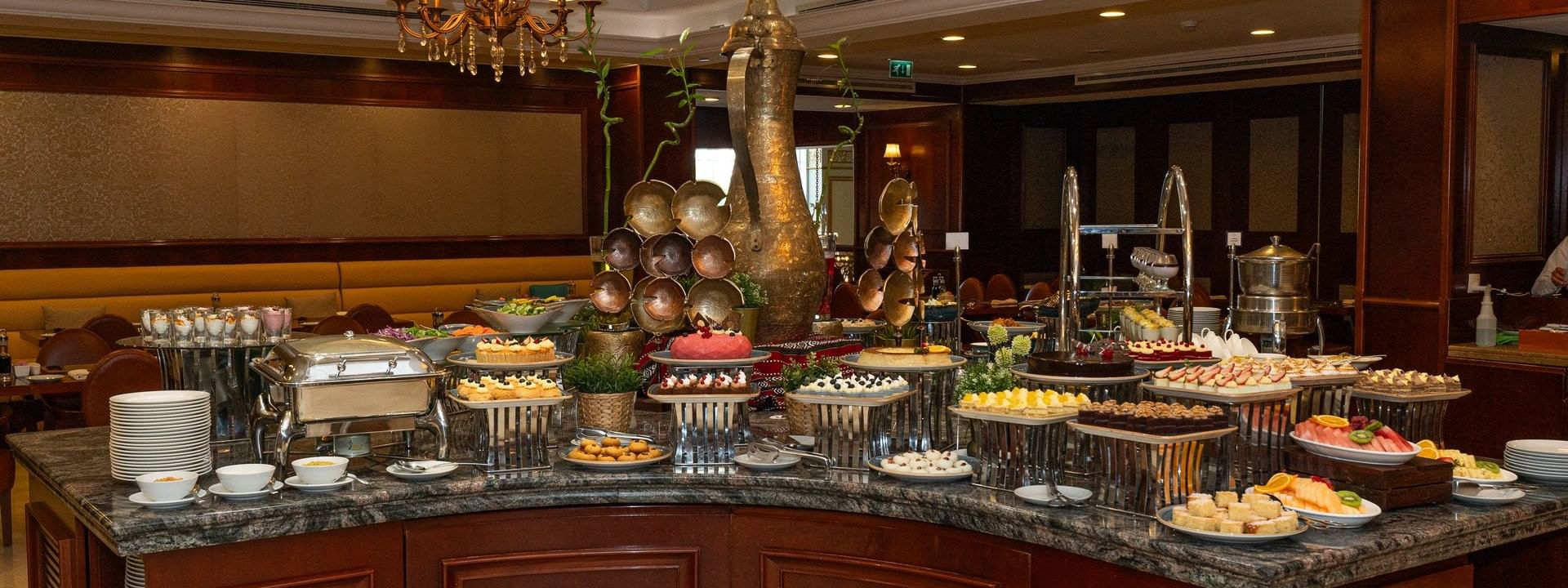 Plated cakes and pastries by a central decorative urn under elegant lighting in L’auberge at Warwick Doha Hotel