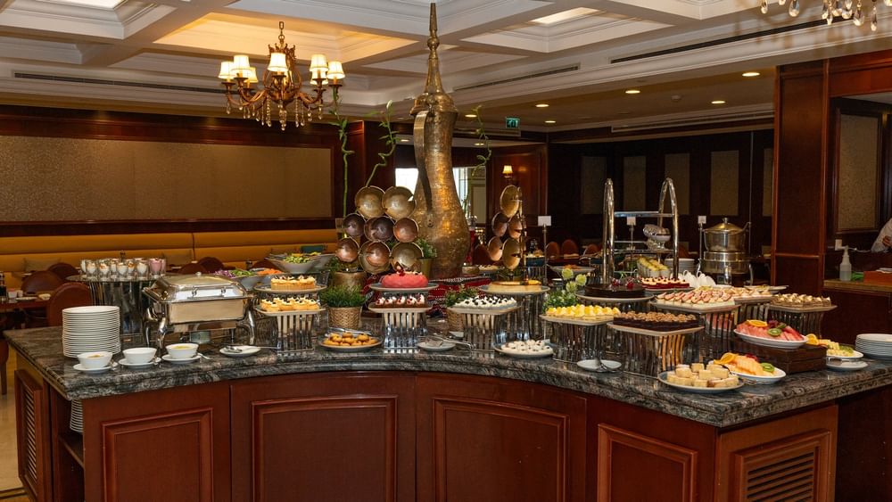 Plated cakes and pastries by a central decorative urn under elegant lighting in L’auberge at Warwick Doha Hotel