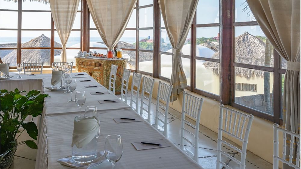 La Palapa venue long table with white chairs by large windows under draped curtains at Quinta Real Huatulco