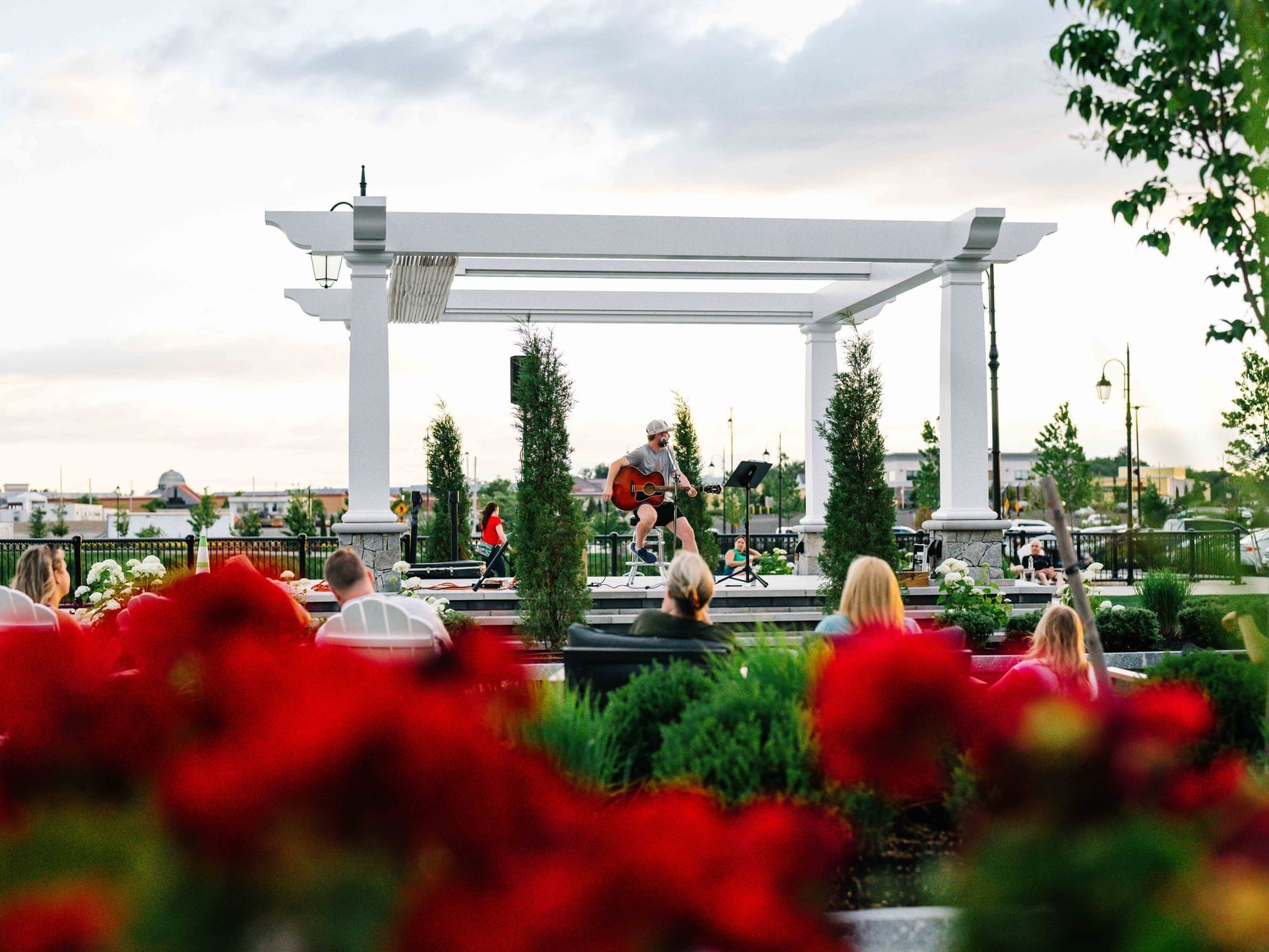 Man plays guitar on stage under a white pergola as people sit around a garden with red flowers.