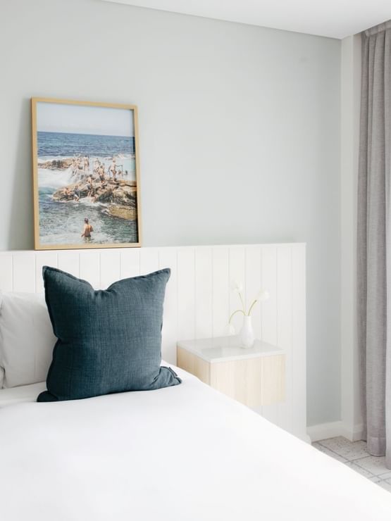 Village Balcony Queen at Elysium Manly with dark teal pillows on a bed by a small nightstand under coastal scenery wall art