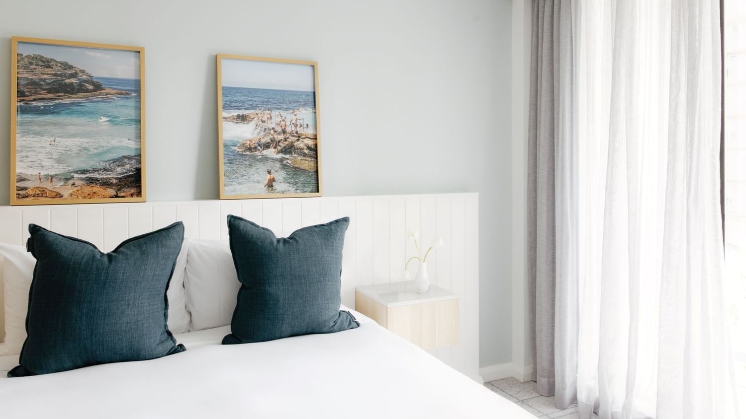 Village Balcony Queen at Elysium Manly with dark teal pillows on a bed by a small nightstand under coastal scenery wall art