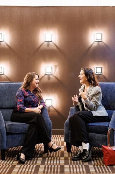 Two professional women chatting in blue armchairs against a modern lit wall in a lounge room at the Rodd Royalty