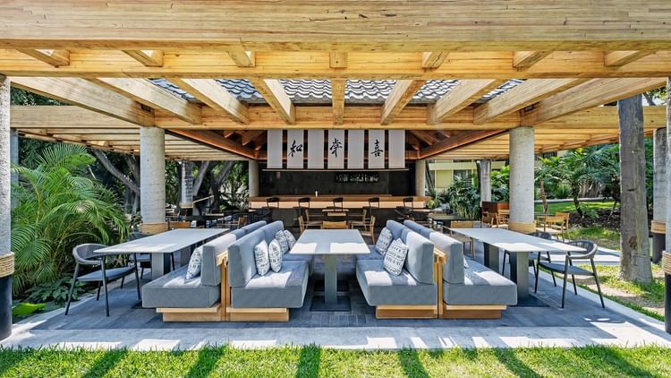 Outdoor dining area with wooden pergola, gray booths, and cushions, surrounded by lush greenery at Grand Fiesta Americana.