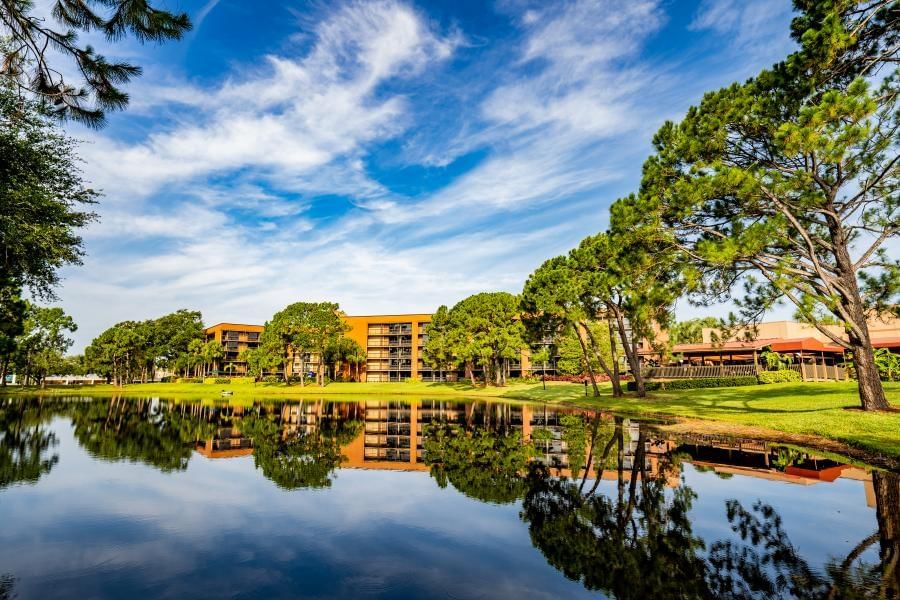 A beautiful reflective lake surrounded by green grass and mature trees with a hotel peeking through in the background. Stay at a Rosen Inn and get all of our helpful tips for visiting Walt Disney World.