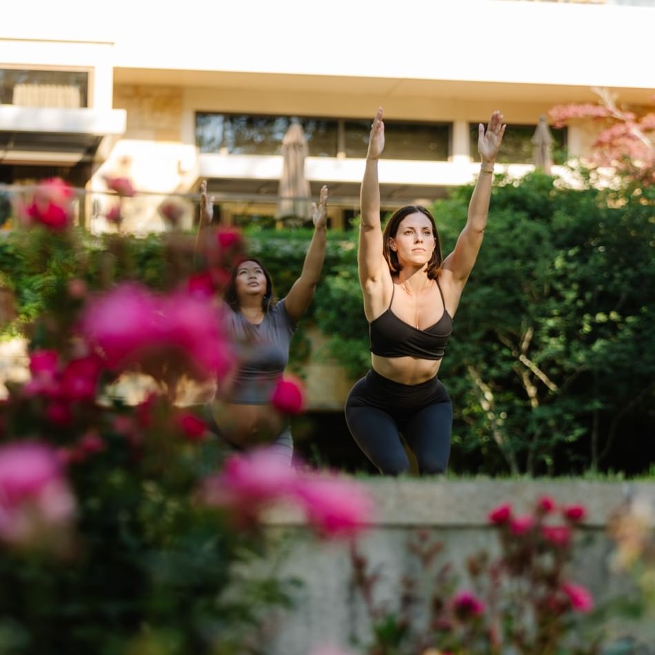 Two women practice yoga with raised arms outdoors surrounded by pink flowers and greenery during Summer Wellness Series.