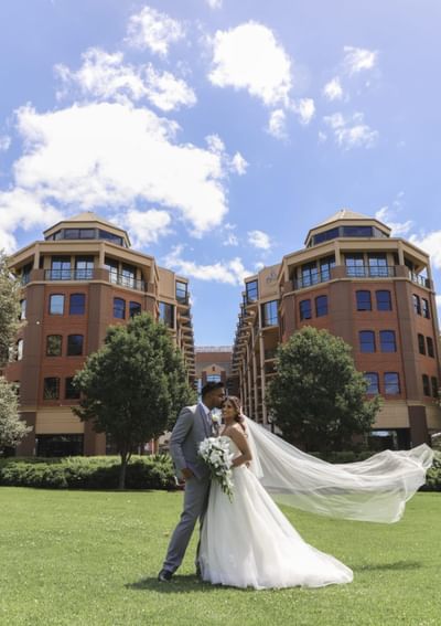 Bride and groom posing closely on a sunny day, her veil flowing in the breeze at Amora Herencia Riverwalk Melbourne