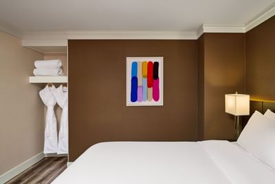 A guest room with a bed, lamp, colorful abstract painting, and a closet with robes and towels.
