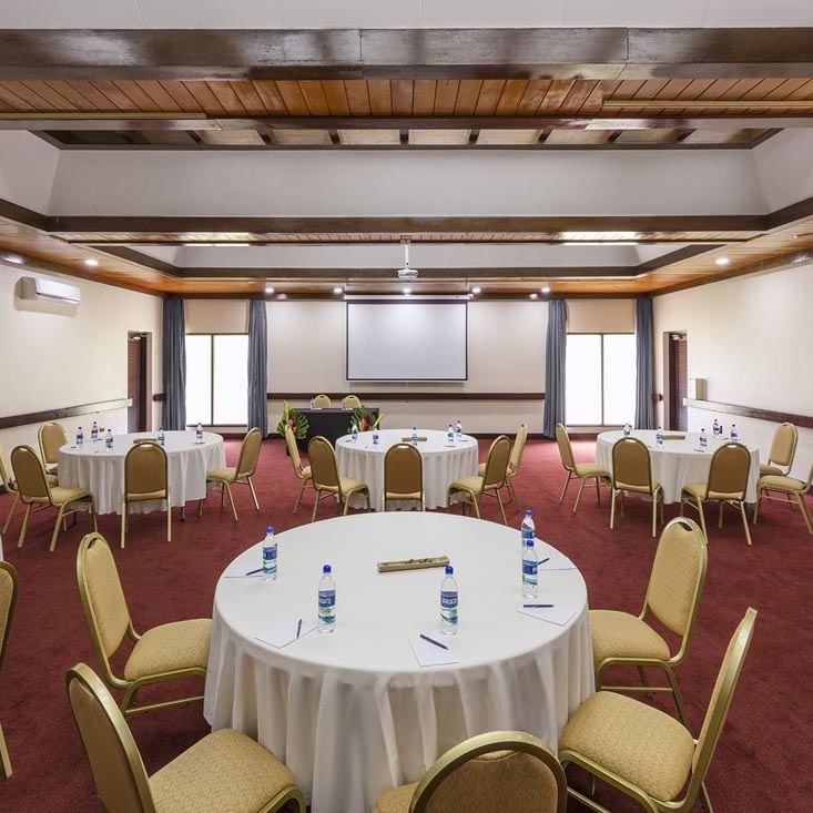 Spacious meeting room with round banquet tables and a projector screen at Warwick Le Lagon