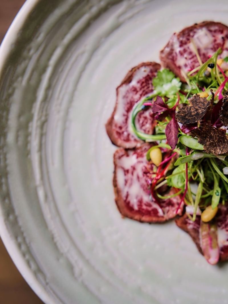 A plate of thinly sliced beef with green vegetables and black truffle toppings