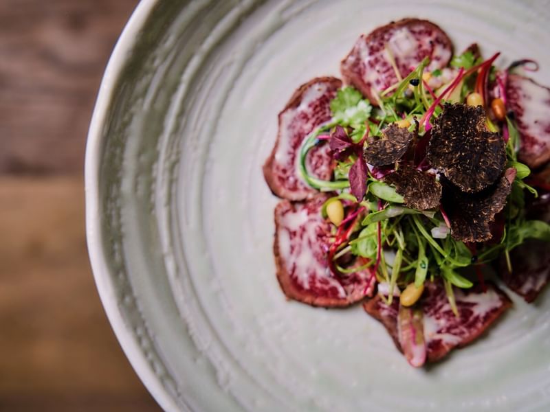 A plate of thinly sliced beef with green vegetables and black truffle toppings
