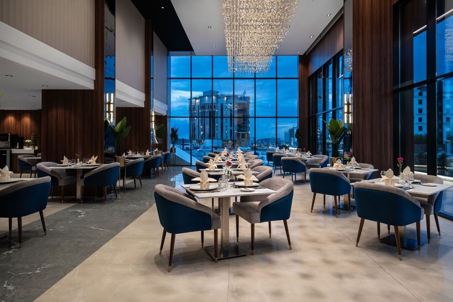 Blue chairs by tables under a crystal chandelier near a window with a city view in Rose Restaurant at Warwick Al Taif Hotel