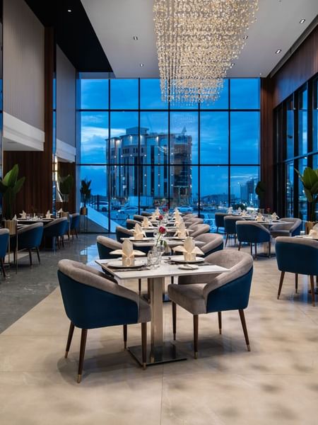 Blue chairs by tables under a crystal chandelier near a window with a city view in Rose Restaurant at Warwick Al Taif Hotel