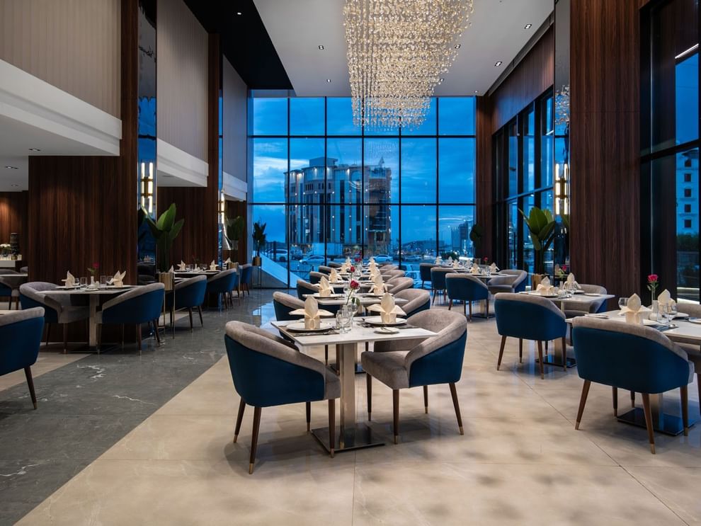 Blue chairs by tables under a crystal chandelier near a window with a city view in Rose Restaurant at Warwick Al Taif Hotel
