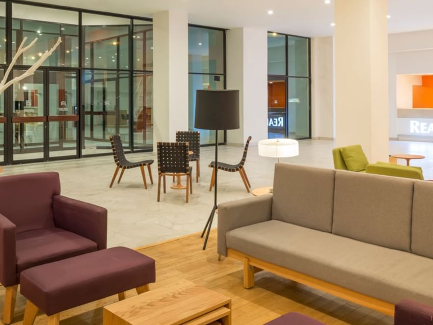 Modern lobby lounge at Real Inn Mexicali, featuring a grey sofa, plum armchairs, and mid-century modern furniture