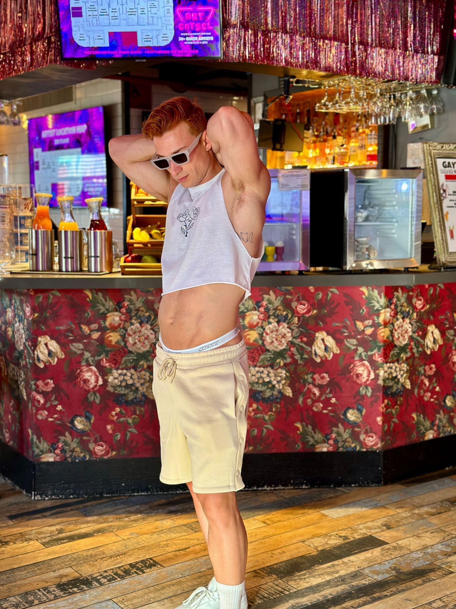 A man standing in front of a bar counter with floral wallpaper and a TV screen.