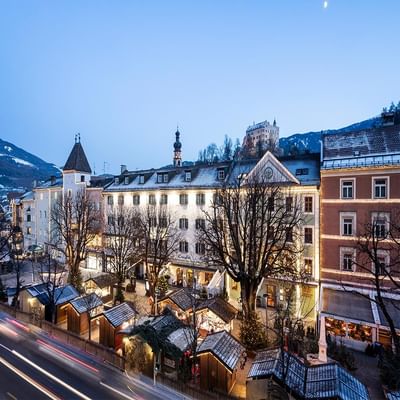 Abendliche Stadtansicht mit beleuchteten Gebäuden, Weihnachtsmarkt und schneebedeckten Bergen im Hintergrund.