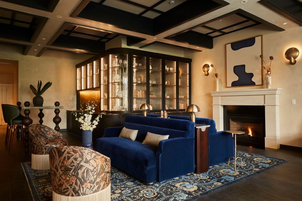 Cozy lounge area featuring plush blue velvet sofas, a warm fireplace, and a stylish illuminated bar at El Prado Hotel