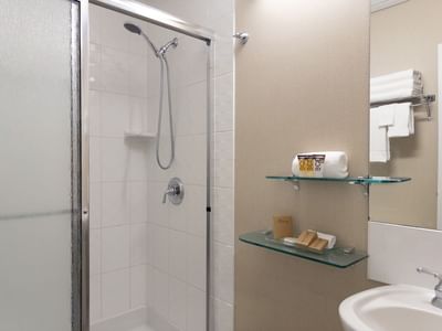 Shower with glass door, towels, toiletries on glass shelves, and a sink at Carriage House Hotel & Conference Centre