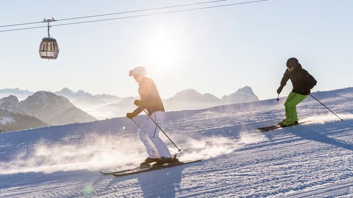 Two skiers navigate a snowy slope with mountains and a ski lift in the background.
