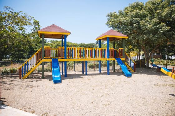 Two colorful playground structures with slides and climbing elements surrounded by trees and a clear blue sky.