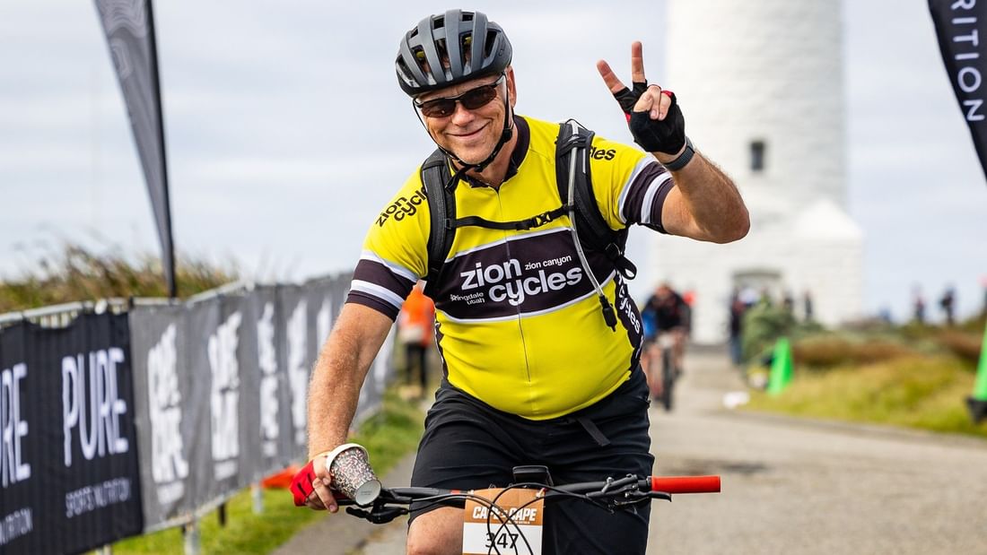 Smiling cyclist in bright yellow jersey gestures peace sign while riding past lighthouse during Cape to Cape MTB 2026.