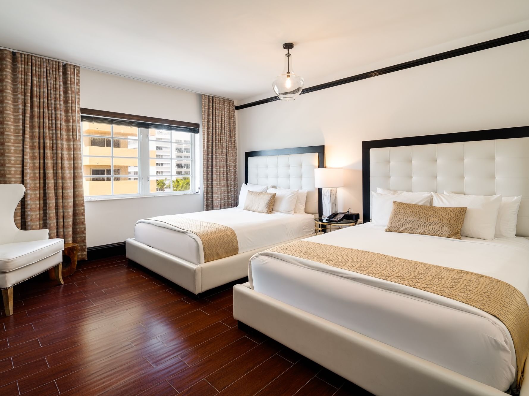 Two beds with pillows, a chair, a window with curtains, and a hanging light fixture in a hotel room.