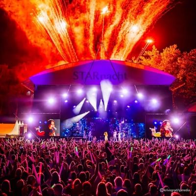 Konzert mit großer Bühne, Scheinwerfern, Feuerwerk und einer Menschenmenge mit Leuchtstäben.
