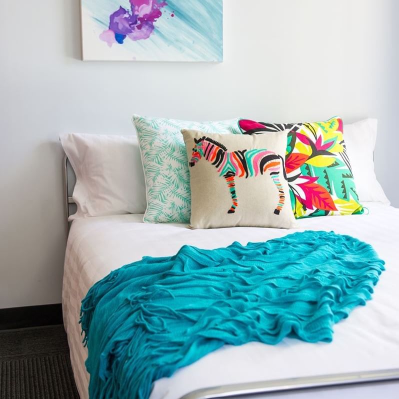 Bright bedroom with white bedding, colorful decorative pillows, turquoise throw, and abstract art at UniLodge on Margaret