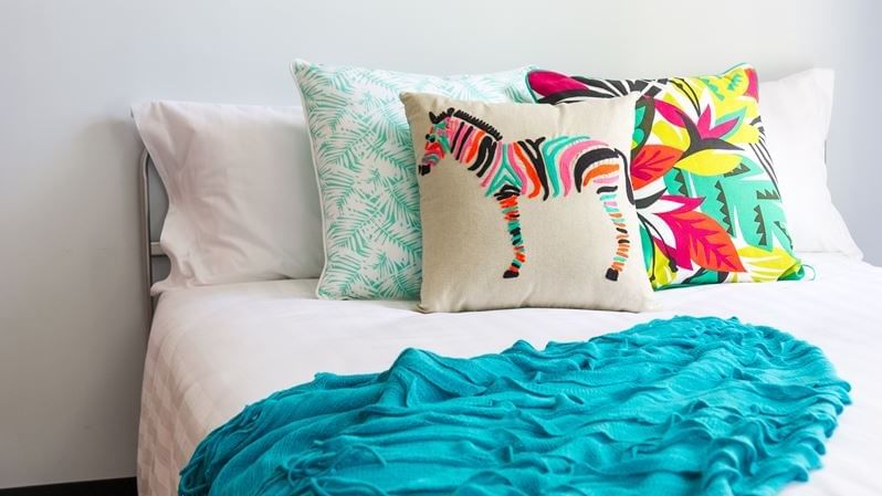 Bright bedroom with white bedding, colorful decorative pillows, turquoise throw, and abstract art at UniLodge on Margaret