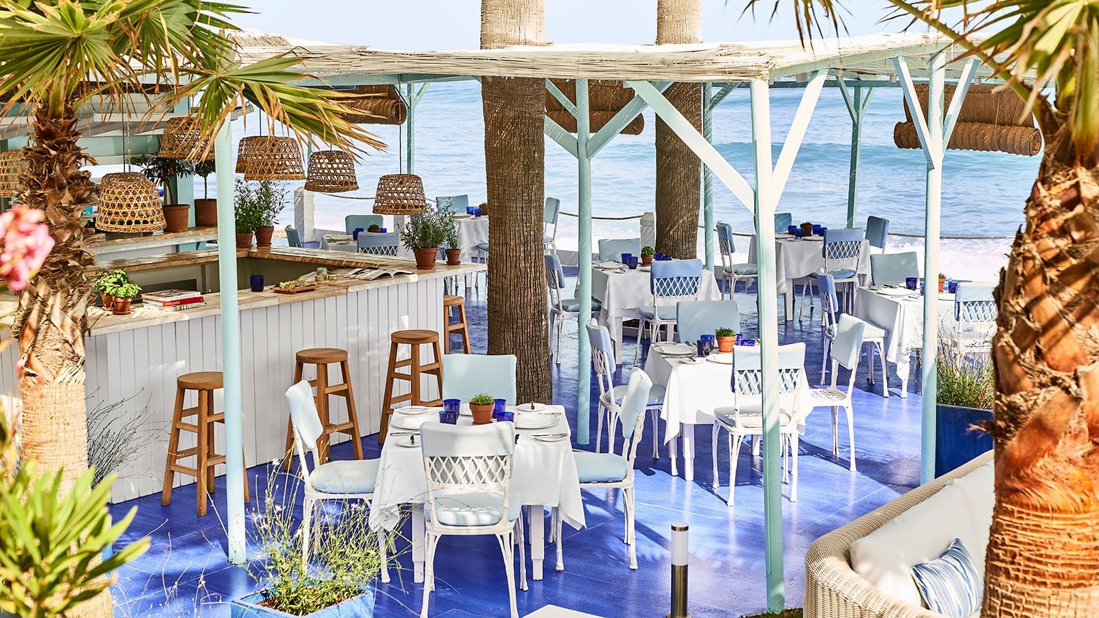 Restaurant with white tables and blue chairs placed under a woven roof by the sea at Marbella Club
