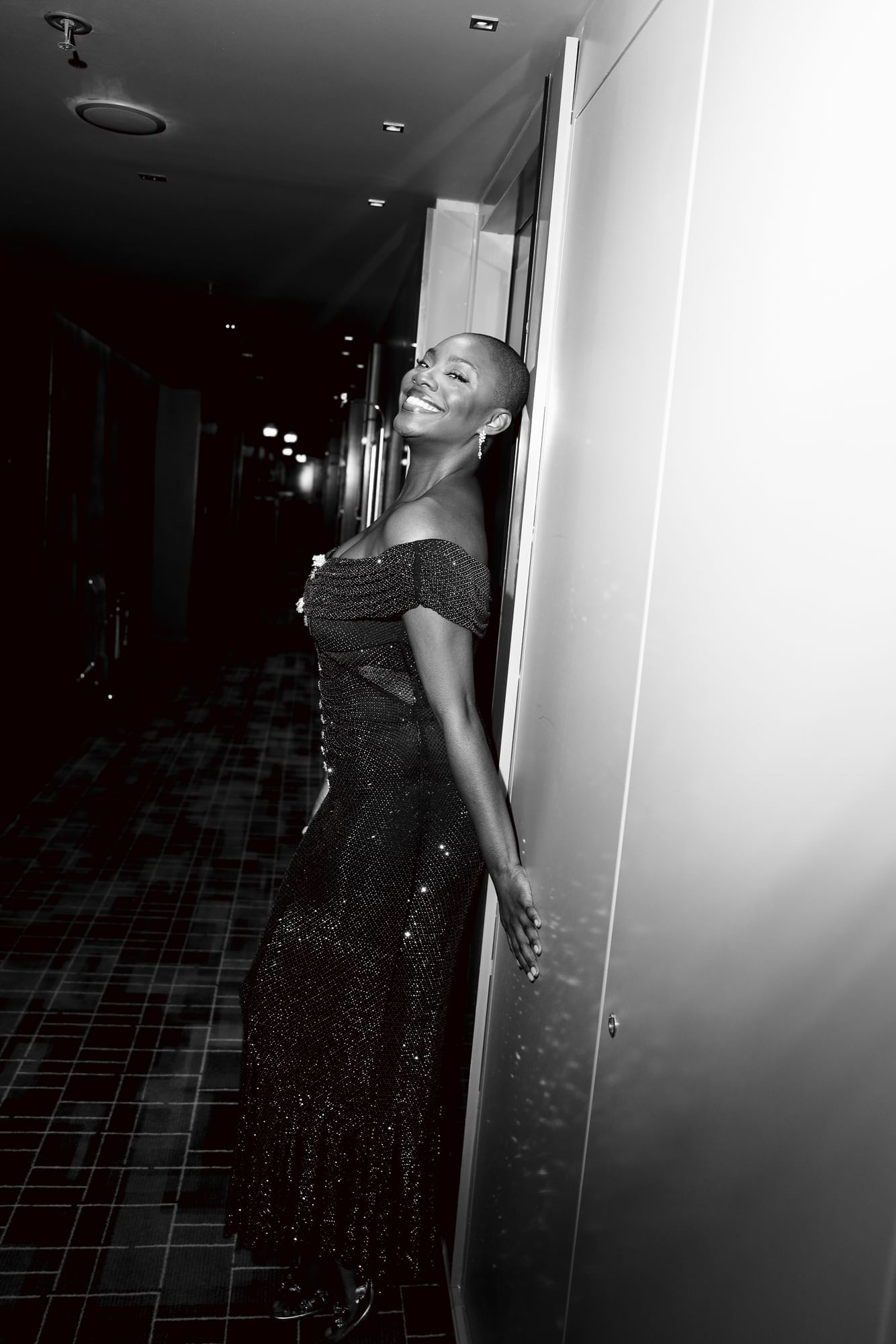 Yomi Adegoke, in a sparkling gown, leans joyfully against a wall in a dimly lit corridor, smiling brightly at The Londoner