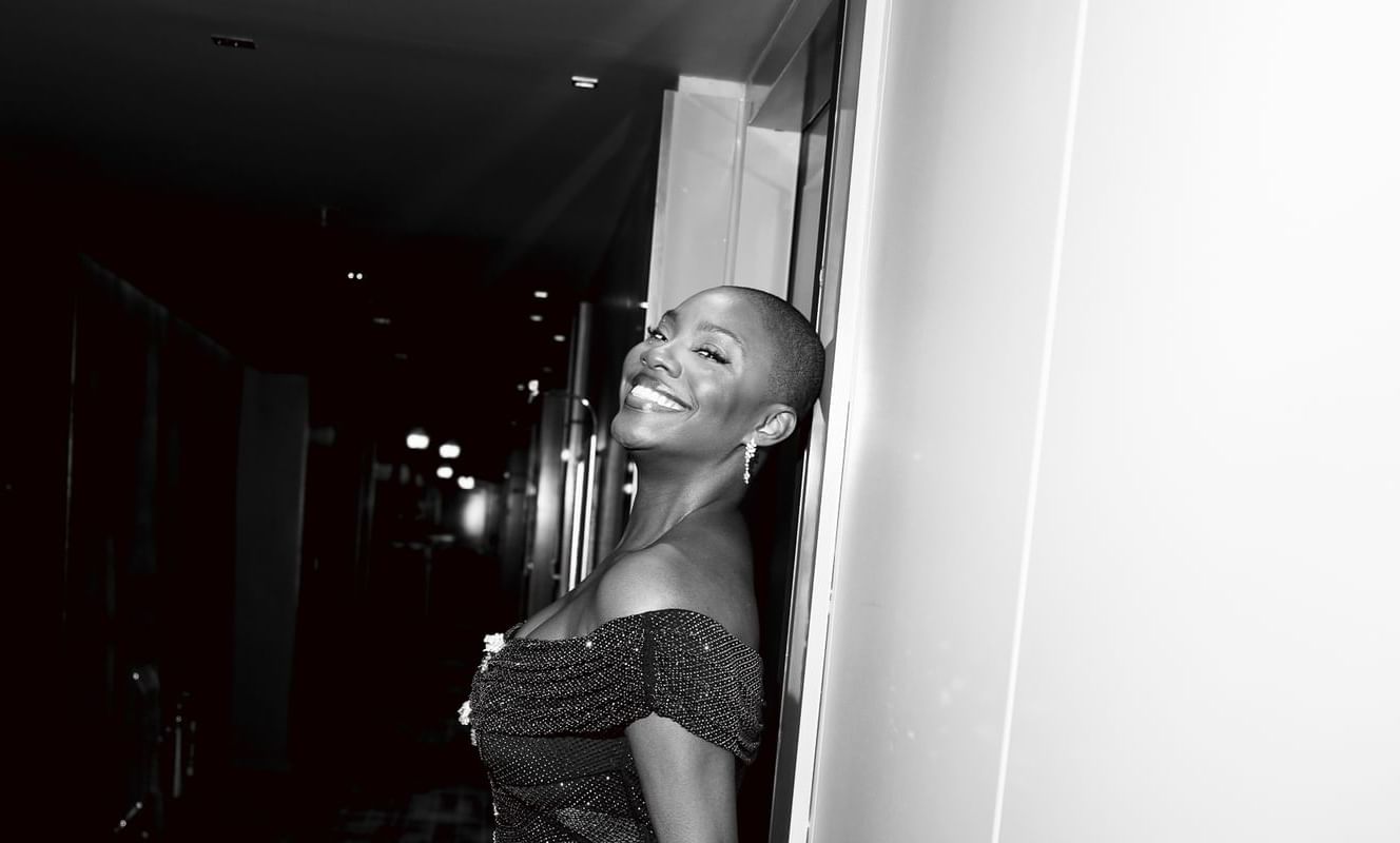 Yomi Adegoke, in a sparkling gown, leans joyfully against a wall in a dimly lit corridor, smiling brightly at The Londoner