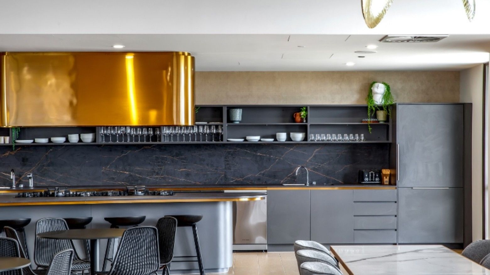 Modern kitchen with marble countertops, gold range hood, gray cabinets, dining area, and a plant on the wall.