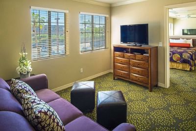 Suite living area with purple sofa, wooden dresser with TV, and windows overlooking the water at London Bridge Resort