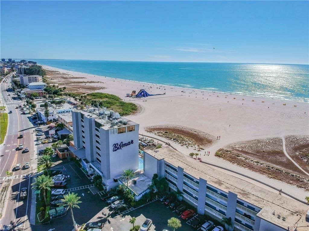 Aerial view of a white beachfront hotel with a parking lot and pool, overlooking a sandy beach and ocean.