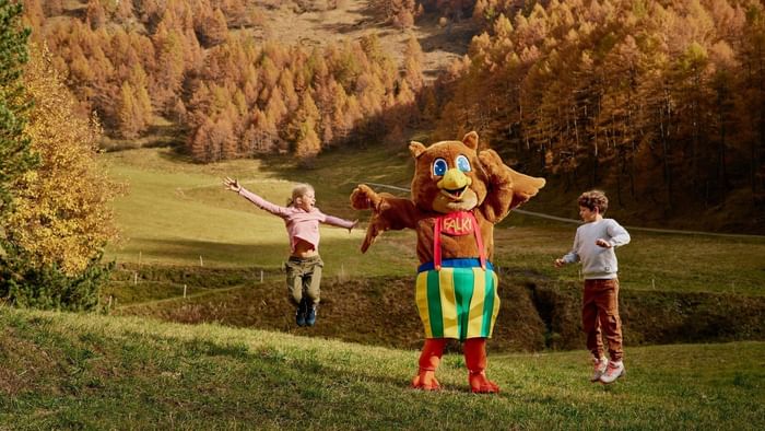 Kids jumping on grass by an owl mascot under autumn trees on a hillside slope near Falkensteiner Hotels & Residences