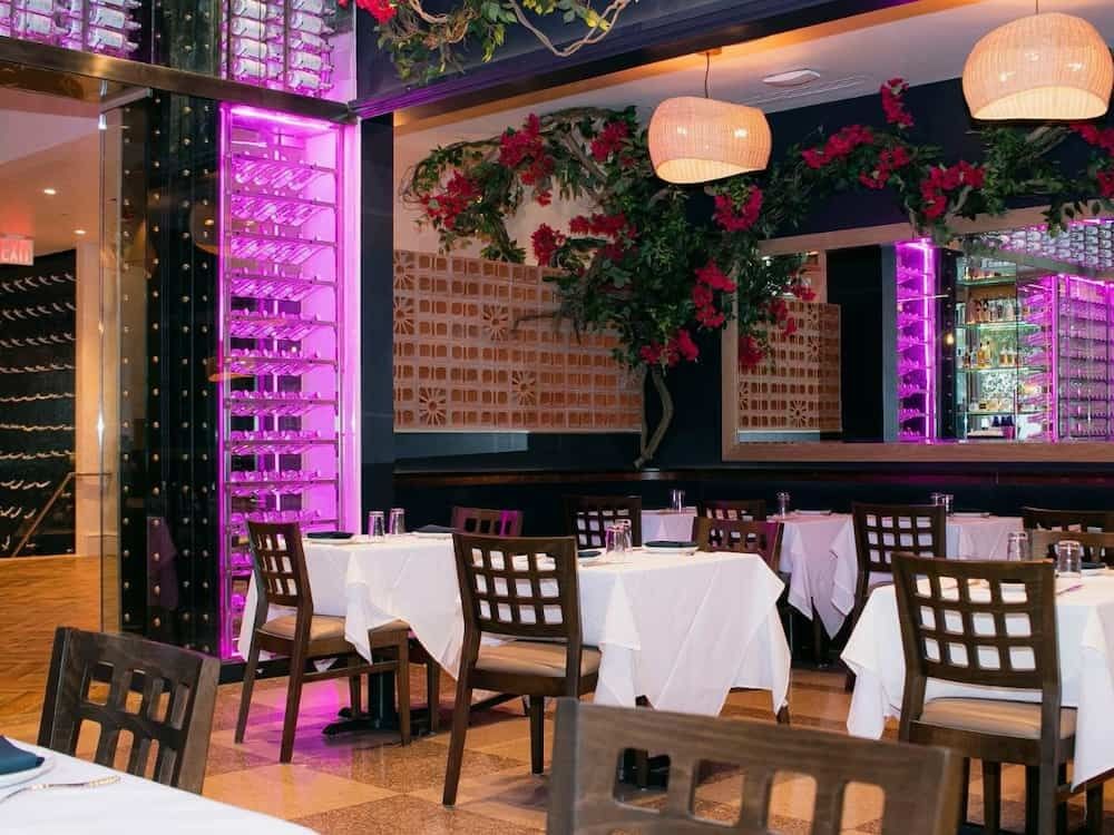 A dining room with white tablecloths, dark wood chairs, and a wine cellar with purple lighting.