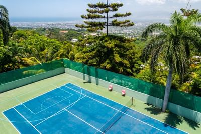 Blue tennis court by red chairs, surrounded by palm trees under a hillside city view at Hotel Montana Haiti