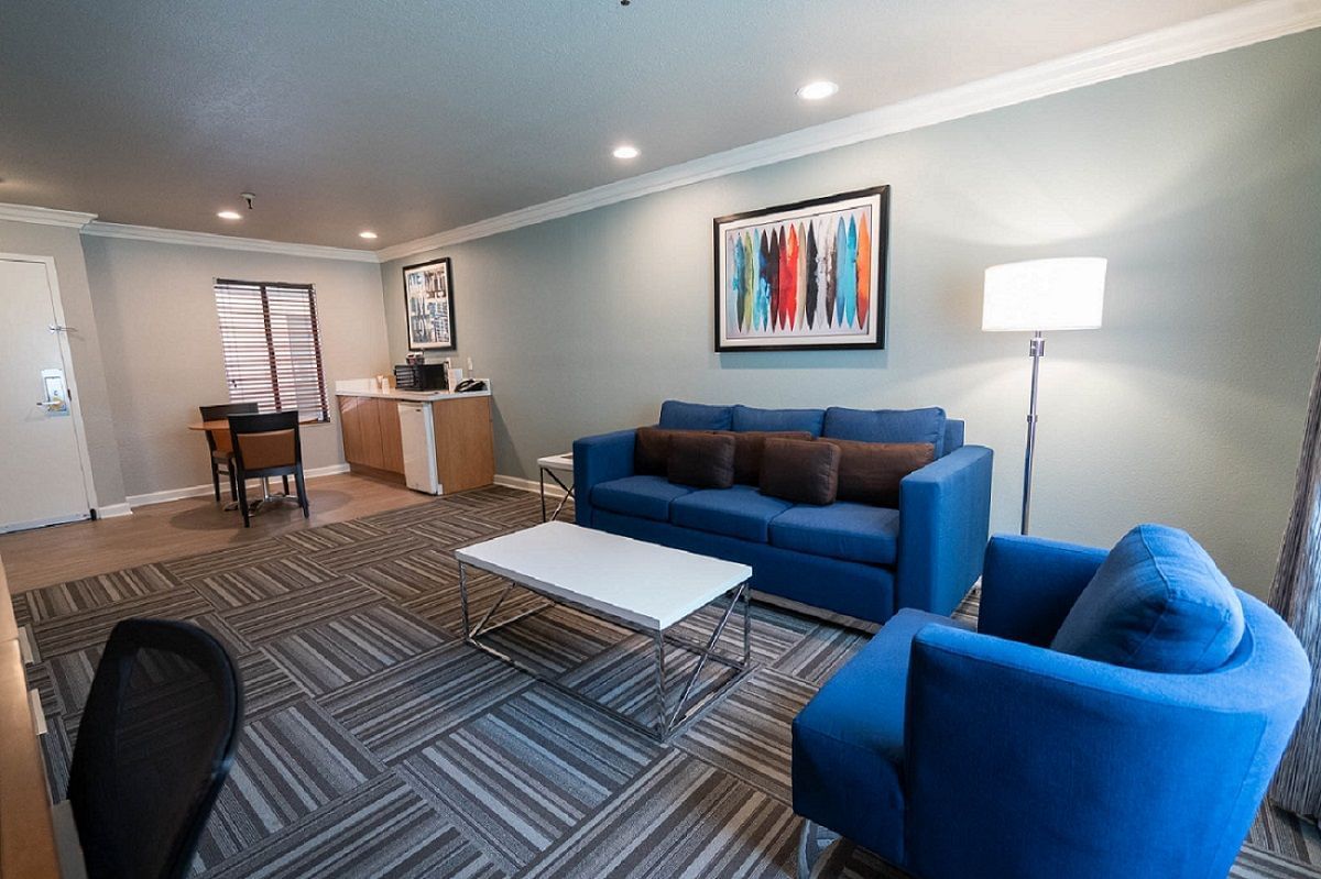 A wide view of a hotel suite's living area with blue sofa and armchair, white coffee table, and abstract art on the wall.