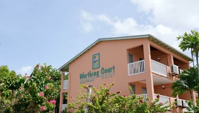 Peach resort building with hotel logo beside green palm trees and bright pink flowers at Worthing Court Apartment