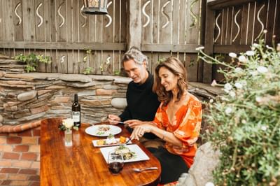 Couple dining outdoors at a wooden table with wine and flowers, near a stone wall and wooden fence.