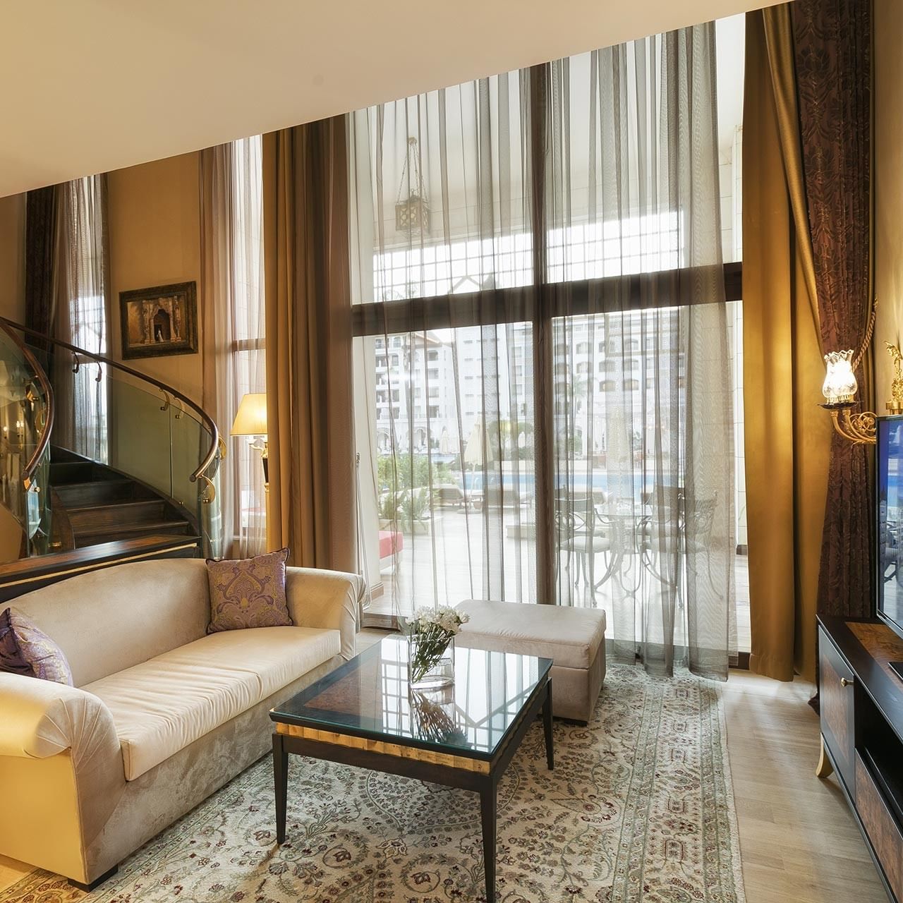 A modern living room with a couch, coffee table, large window, spiral staircase, and flat-screen TV in Pool Duplex Suite at Titanic Mardan Palace.