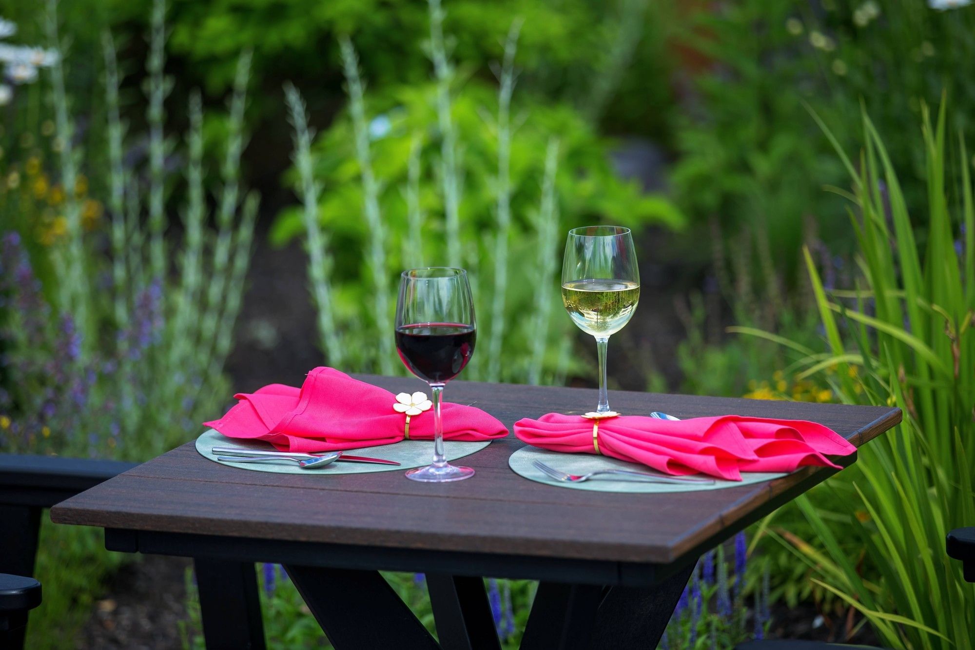 Two wine glasses beside folded bright pink napkins on dark wooden table in lush green garden dining at The Brook