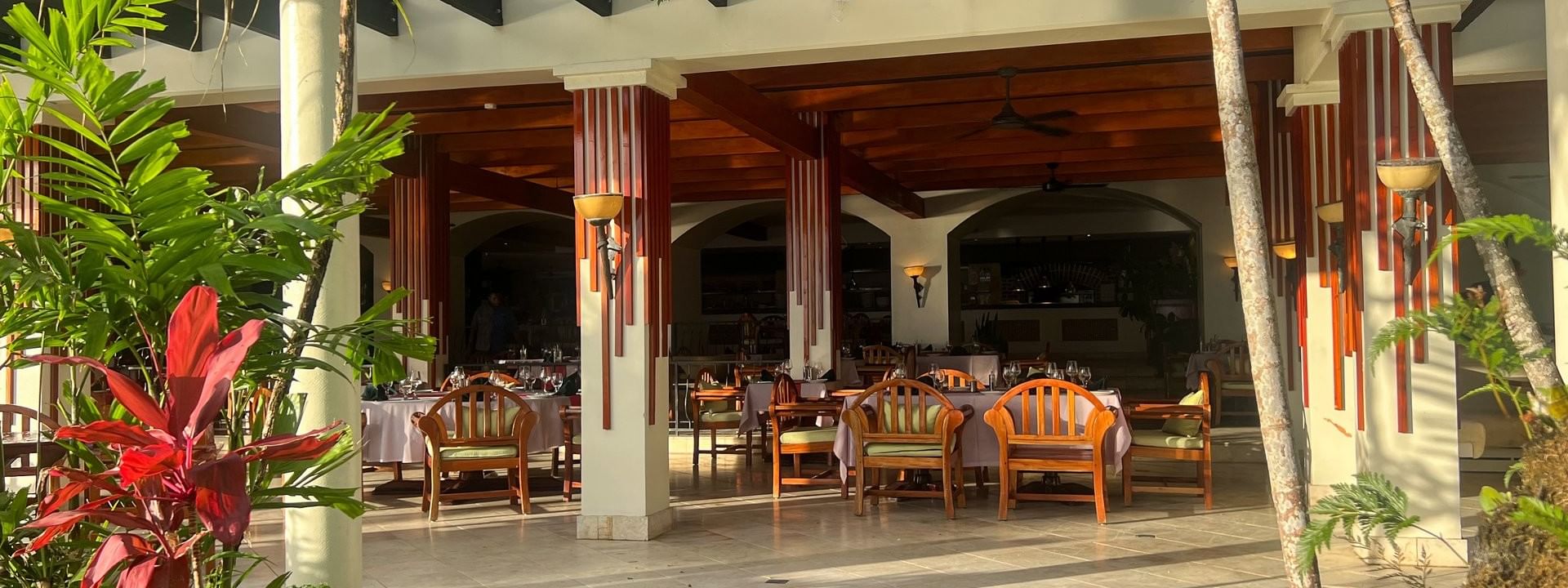 Wooden chairs by white tables under a dark pergola near vibrant red leaves at in Pappagallo Warwick Fiji Resort and Spa