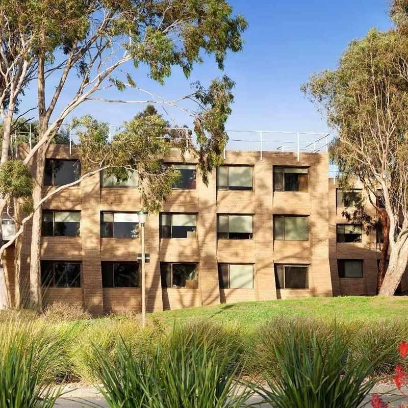 A large brick building with multiple windows surrounded by tall trees and greenery.