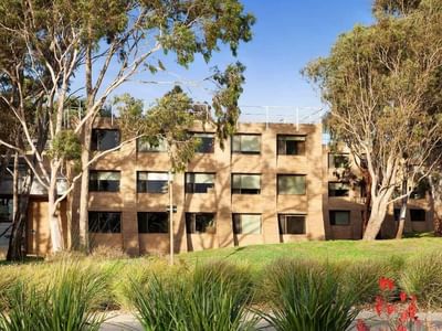 A large brick building with multiple windows surrounded by tall trees and greenery.