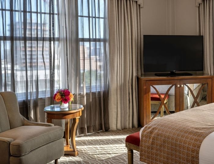Flat-screen TV on a wooden stand by a chair under drapes in Corner King at Warwick Melrose Dallas